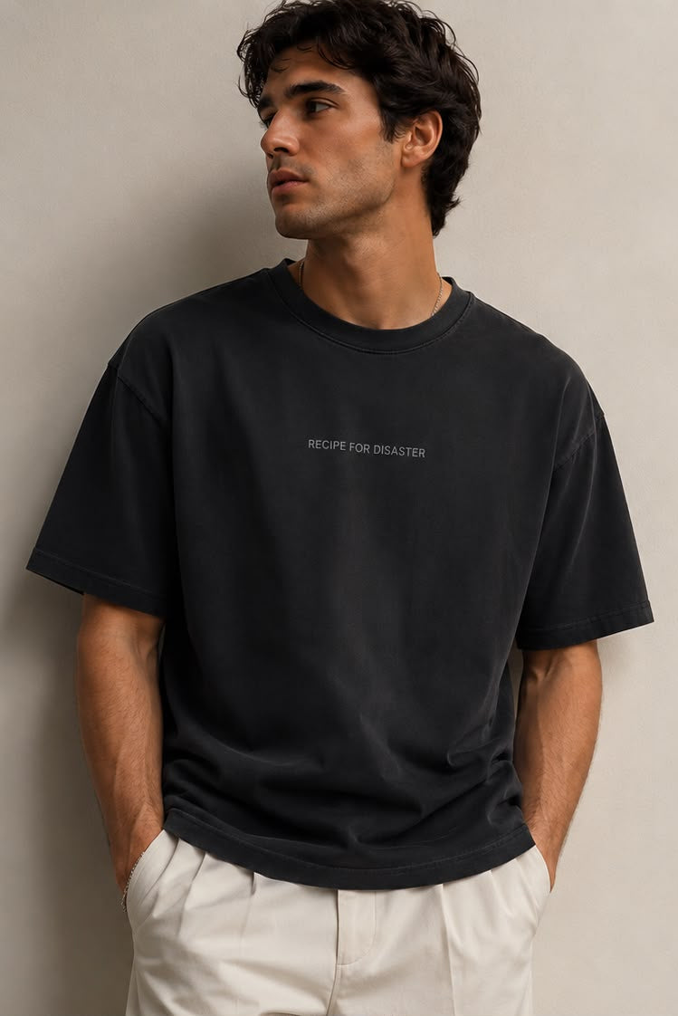 A man wearing an oversized black short-sleeve t-shirt with 'RECIPE FOR DISASTER' printed in white on the chest.